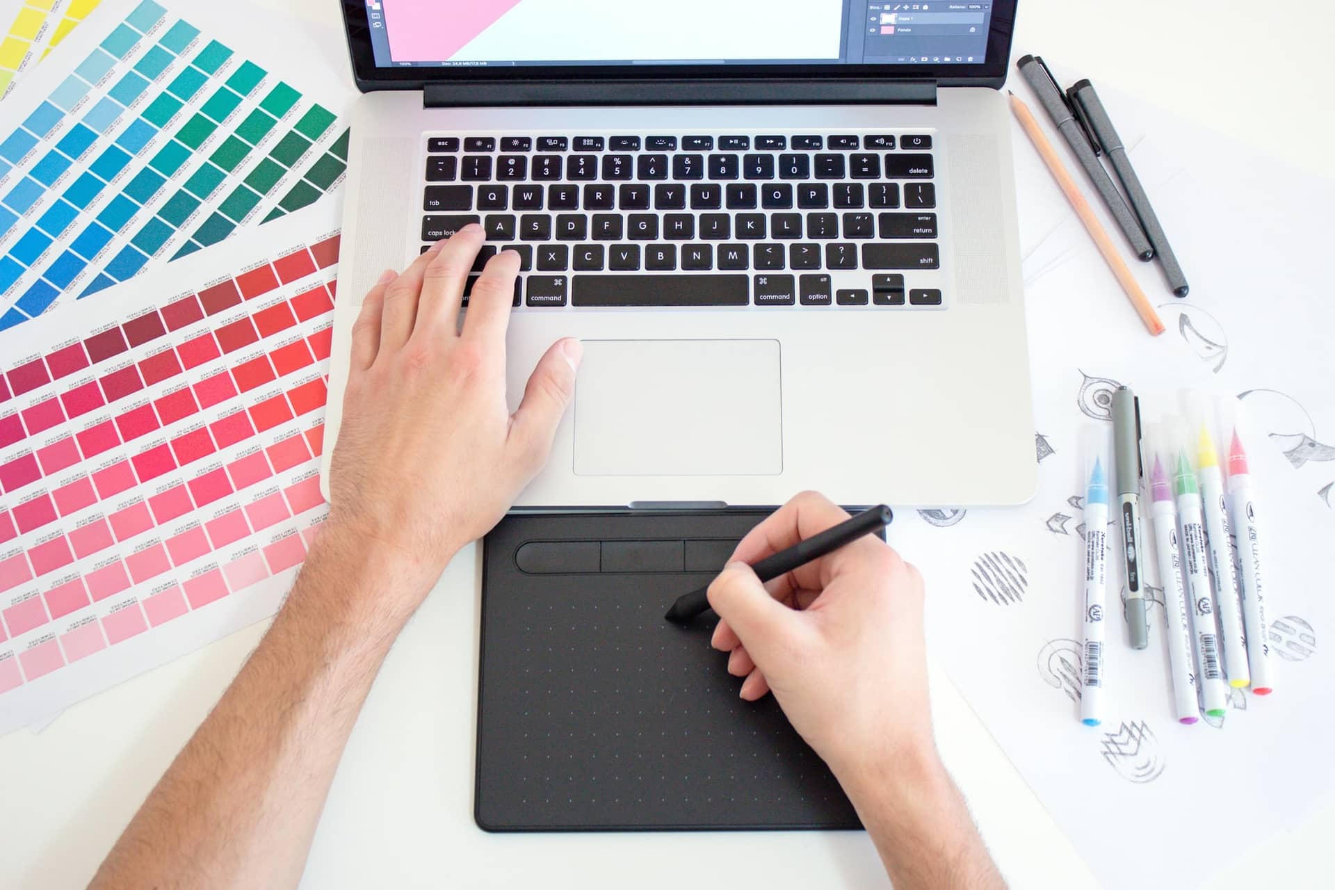 Overhead view of a designer's hands on a laptop and a pen tablet, beside colour swatch sheets, brush markers and sketched icons on paper.