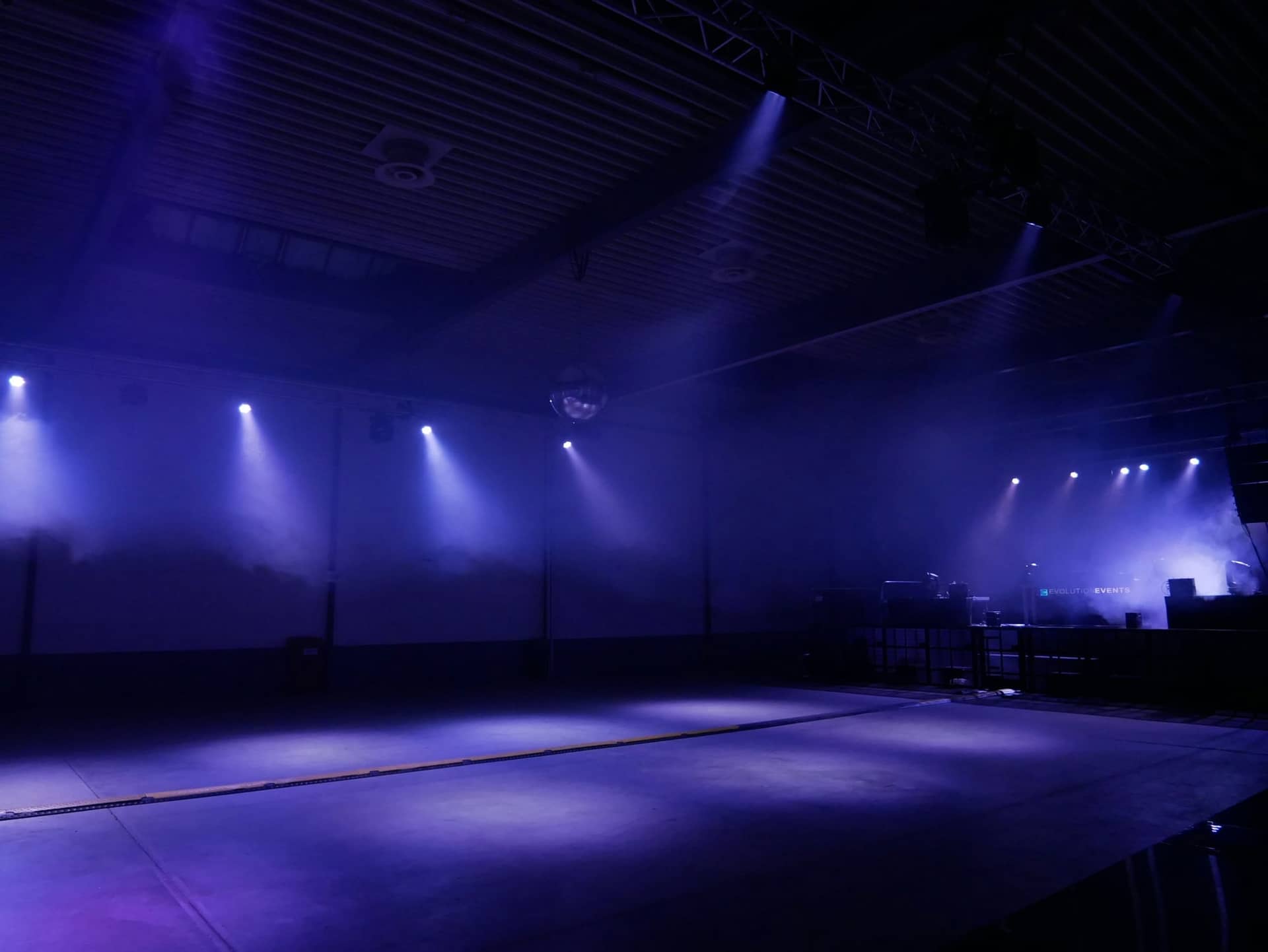 An empty event hall lit in blue and violet, spotlight beams cutting through haze from a lighting truss towards the stage.