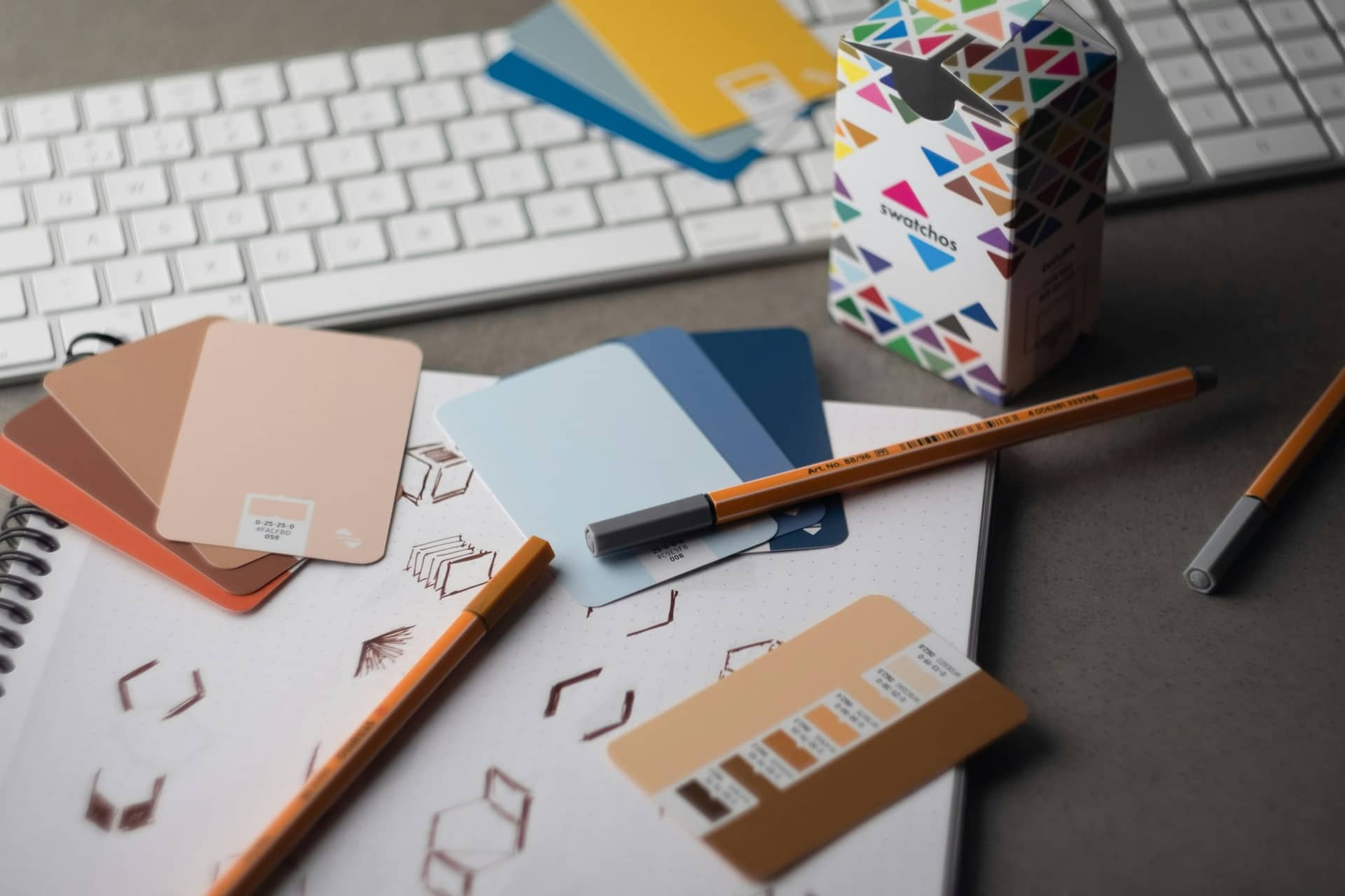 Colour swatch cards, orange fineliner pens and a dot-grid sketchbook with hand-drawn shapes on a grey desk in front of a white keyboard.