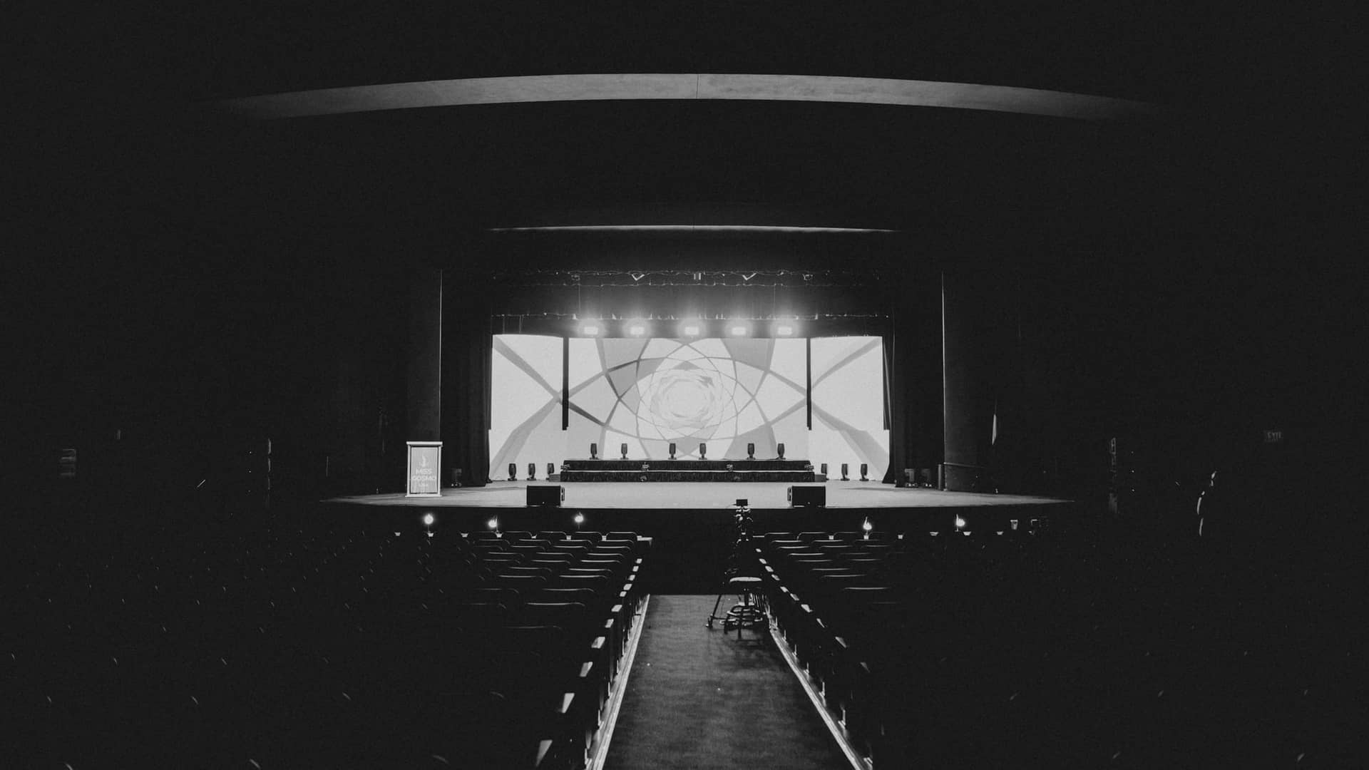Black-and-white auditorium seen from the back of the hall: a lit stage with a geometric backdrop at the far end, rows of seats in shadow either side of the centre aisle.