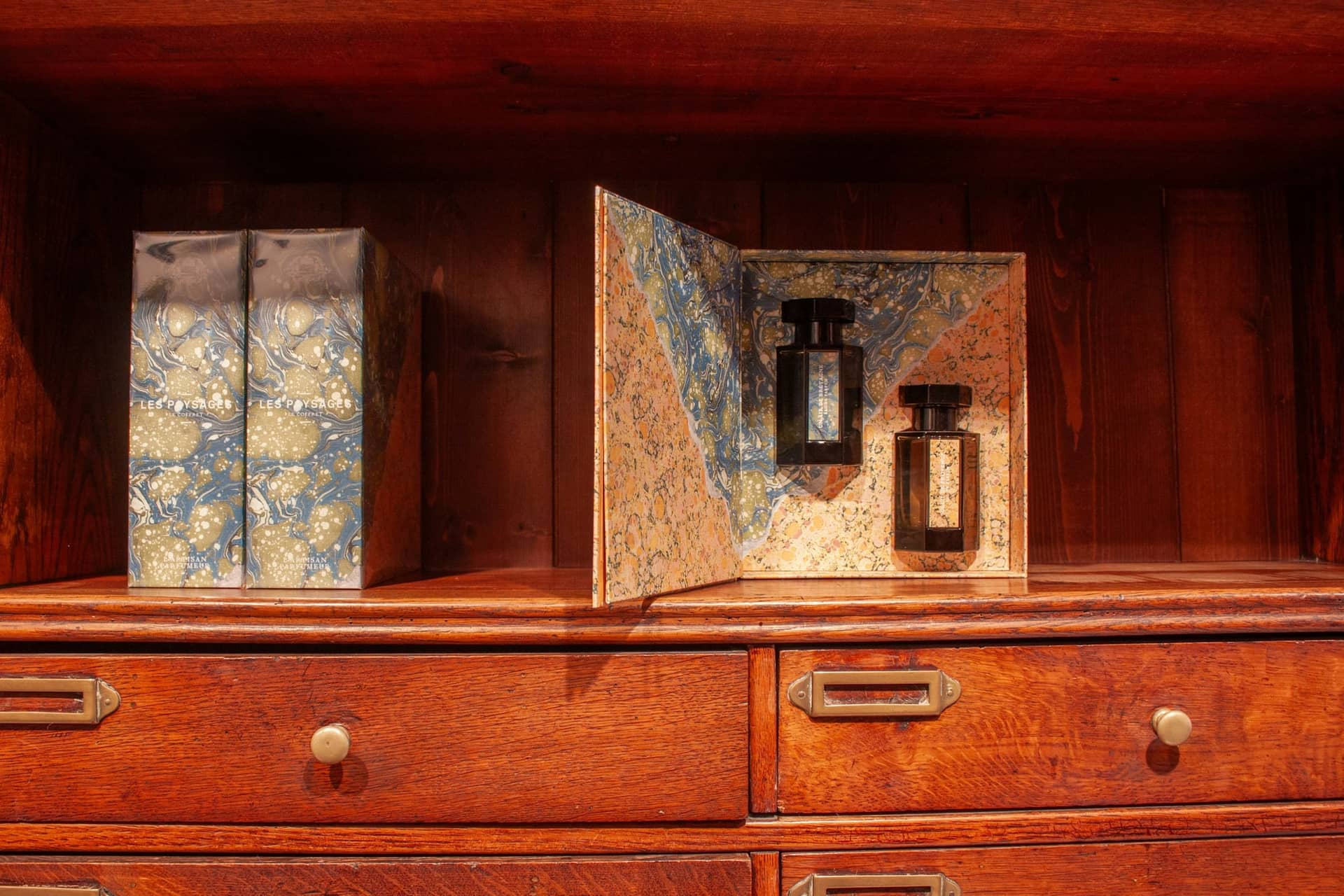 Marbled-paper fragrance boxes on a wooden shelf, one presentation box standing open to show two perfume bottles inside.
