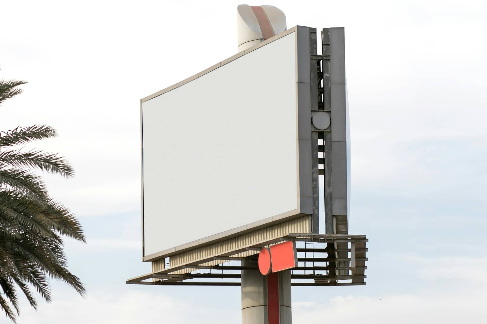 A large blank roadside billboard on a tall steel pole against a pale sky, palm fronds at the left edge.