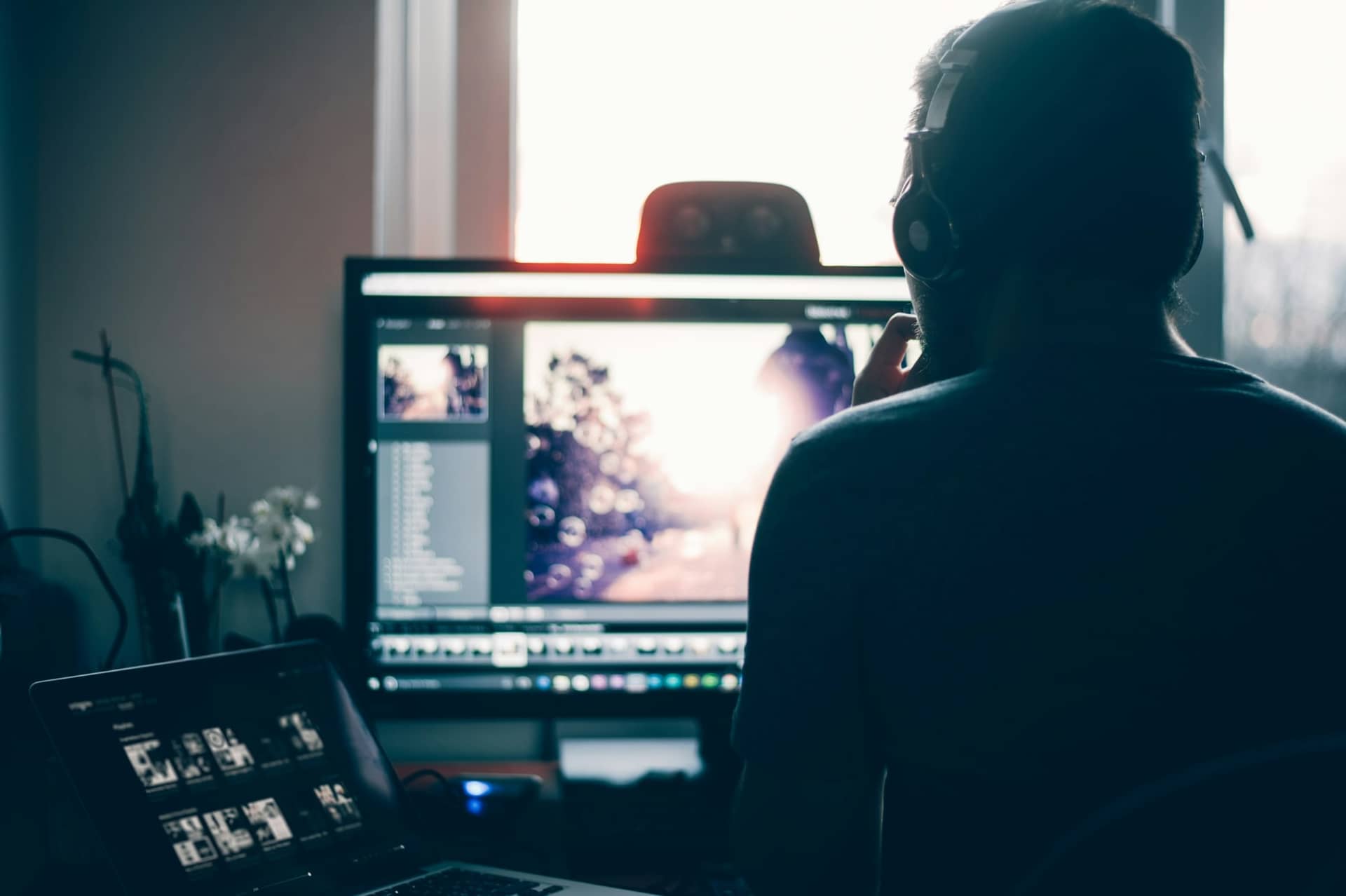 A designer seen from behind, backlit by a window, editing a photograph on a large monitor with a laptop beside it.