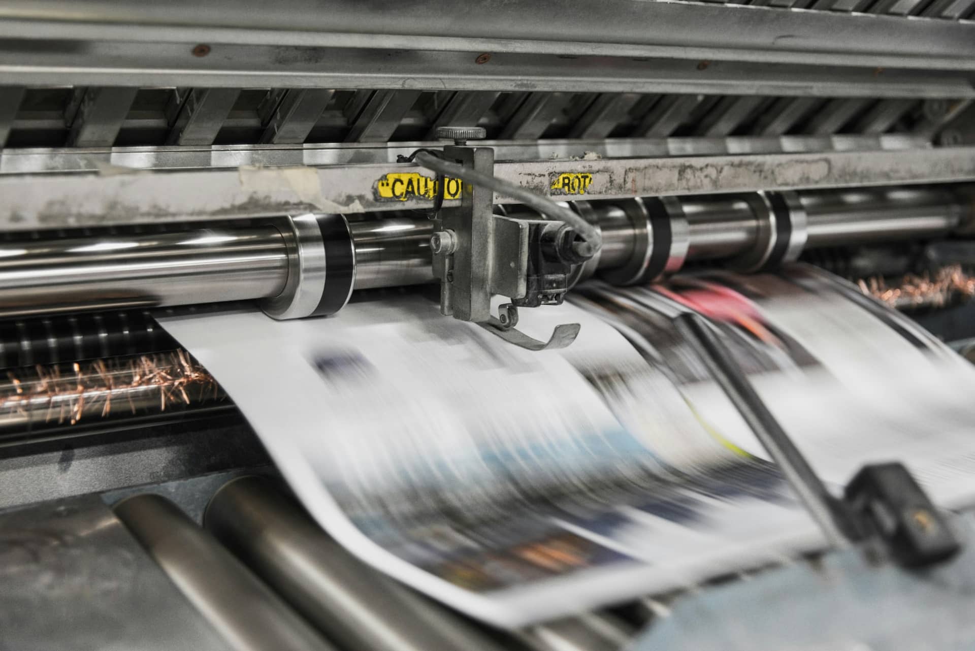 Close-up of a printing press: freshly printed sheets running at speed between steel rollers, blurred by motion.